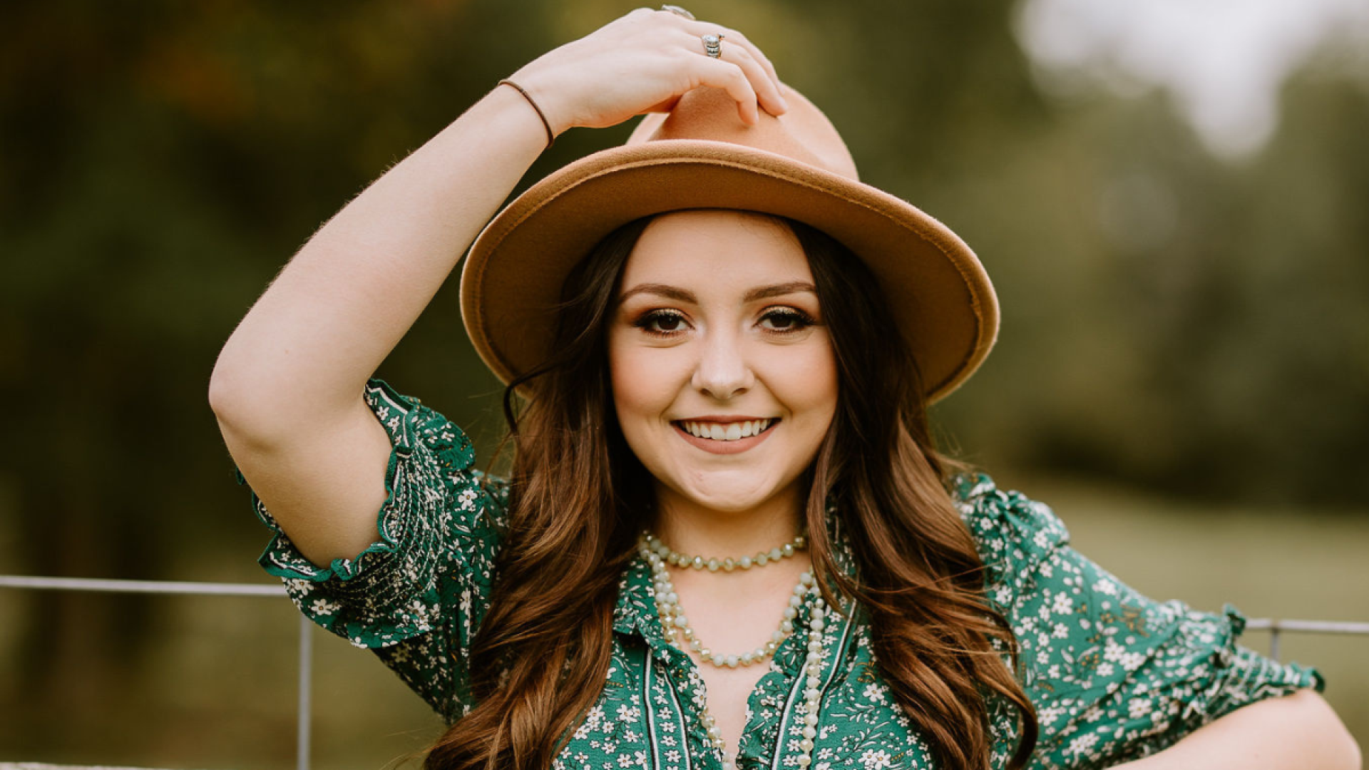 A young woman in a green floral top and brown wide-brimmed hat smiles at the camera outdoors, one hand resting on top of the hat.